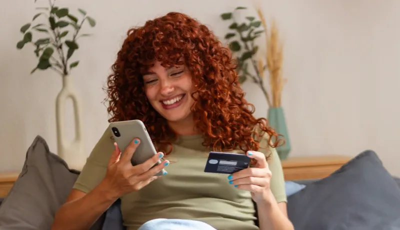 Une femme souriante utilisant son smartphone et tenant une carte bancaire pour un achat en ligne.