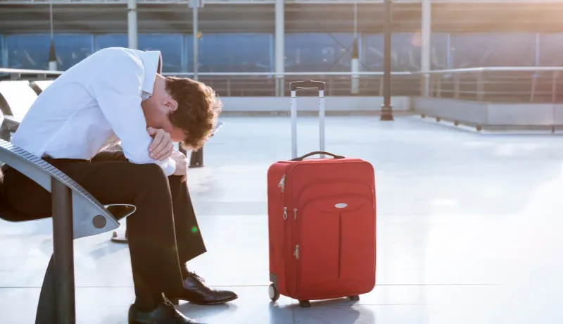 Un homme accablé assis dans un aéroport à côté d'une valise rouge, illustrant un retard de vol.