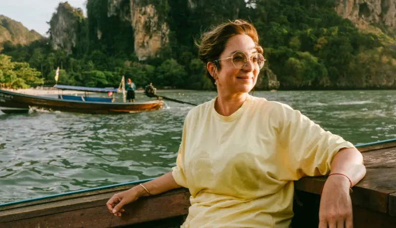 Femme souriante avec lunettes de soleil sur un bateau traditionnel en mer, avec des falaises tropicales en arrière-plan.