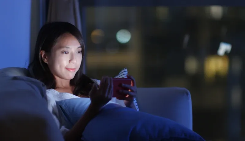 Jeune femme dans la pénombre, le visage éclairé par l'écran de son smartphone alors qu'elle est allongée sur son canapé.