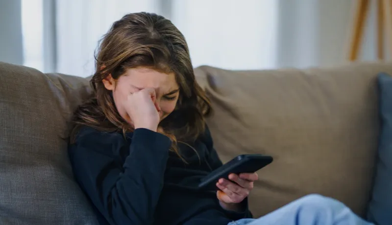 Jeune fille assise sur un canapé, l'air triste et se frottant les yeux en regardant son téléphone portable.