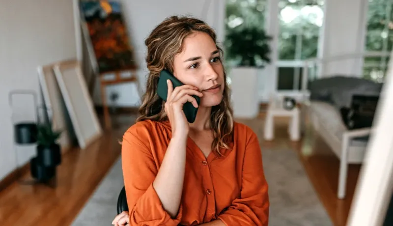 Femme pensive dans un intérieur lumineux, tenant son smartphone contre son oreille lors d'un appel téléphonique.