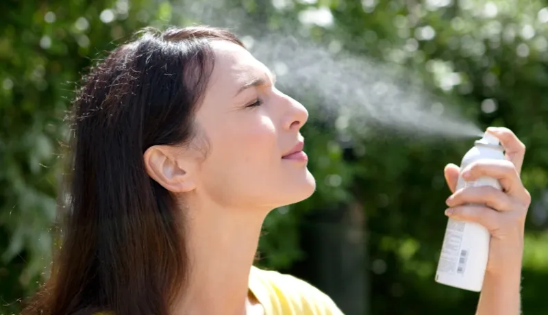 Femme fermant les yeux et s'aspergeant le visage avec un brumisateur d'eau en extérieur par temps ensoleillé.