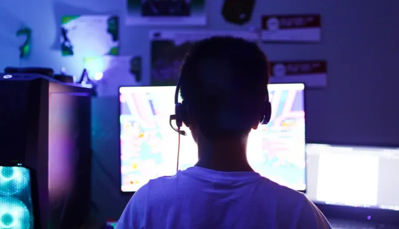 Vue de dos d'une personne avec un casque audio devant plusieurs écrans d'ordinateur dans une pièce sombre éclairée par des LED bleues.