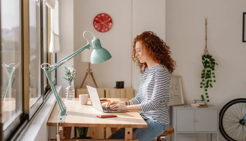 Femme aux cheveux bouclés souriant tout en travaillant sur son ordinateur portable dans un bureau lumineux et moderne.