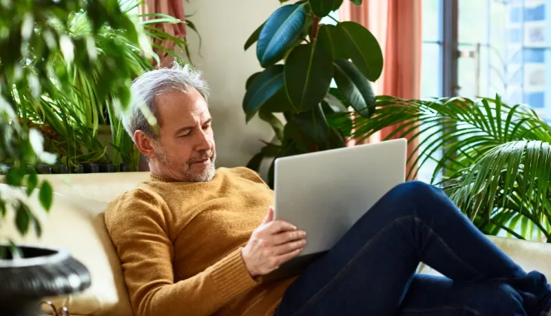 Homme mûr assis confortablement sur un canapé entouré de plantes vertes, consultant attentivement son ordinateur portable.