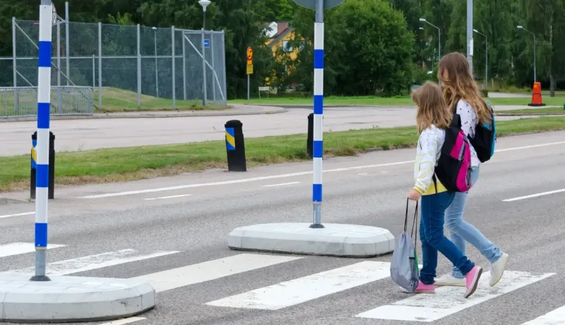 Deux écolières avec leurs sacs à dos traversent prudemment un passage piéton sécurisé par des poteaux bleus.