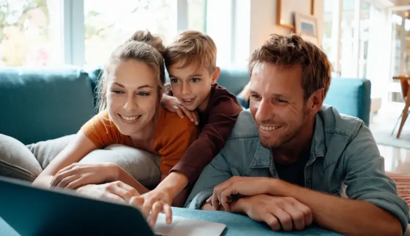 Un couple et leur jeune fils regardent ensemble un ordinateur portable avec le sourire sur un canapé bleu.
