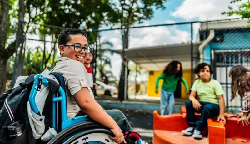 Un garçon souriant à lunettes en fauteuil roulant s'amuse avec ses camarades dans une cour de récréation.