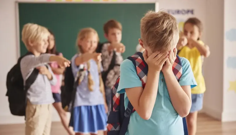 Un jeune garçon triste se cache le visage pendant que d'autres enfants le pointent du doigt en classe.