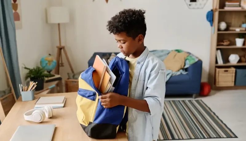 Un jeune garçon aux cheveux bouclés remplit avec sérieux son sac à dos jaune et bleu de plusieurs livres.