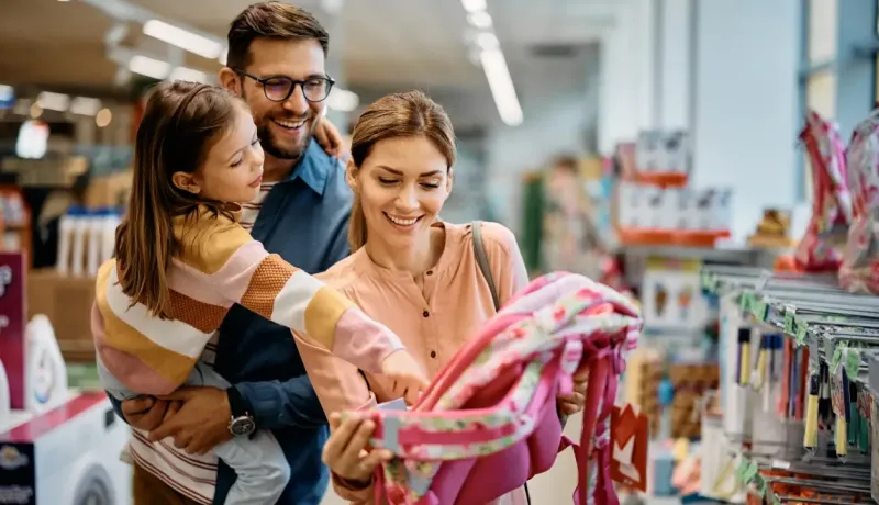 Un couple souriant et leur fille choisissent ensemble un cartable rose dans les rayons d'un magasin.