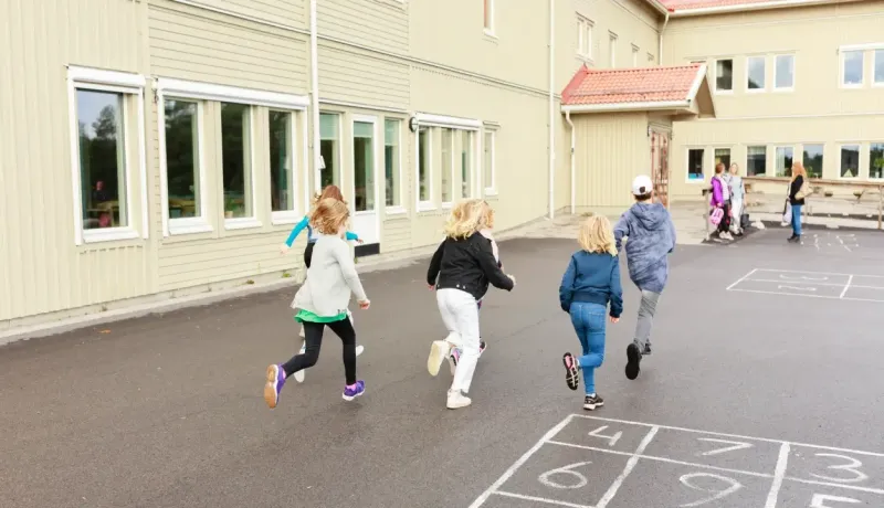 Un groupe d'écoliers court avec enthousiasme dans la cour de récréation vers un bâtiment scolaire moderne.