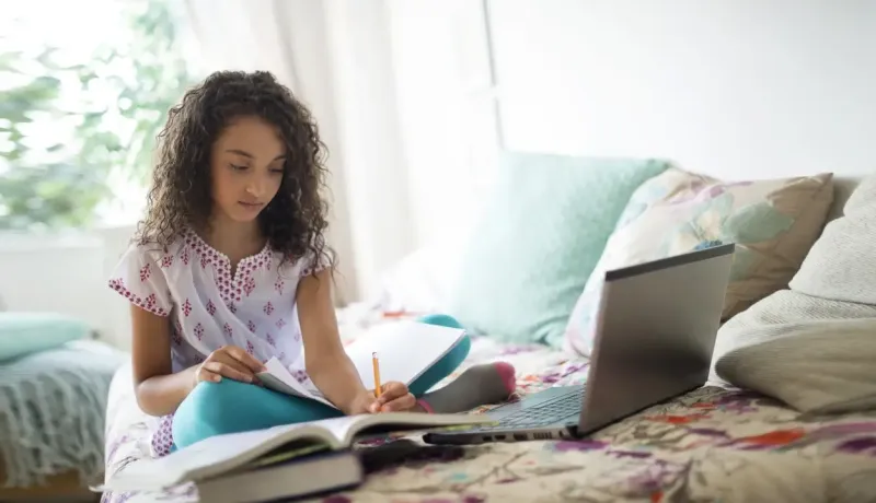Une jeune fille aux cheveux frisés révise attentivement ses leçons sur son lit avec un ordinateur et des cahiers.