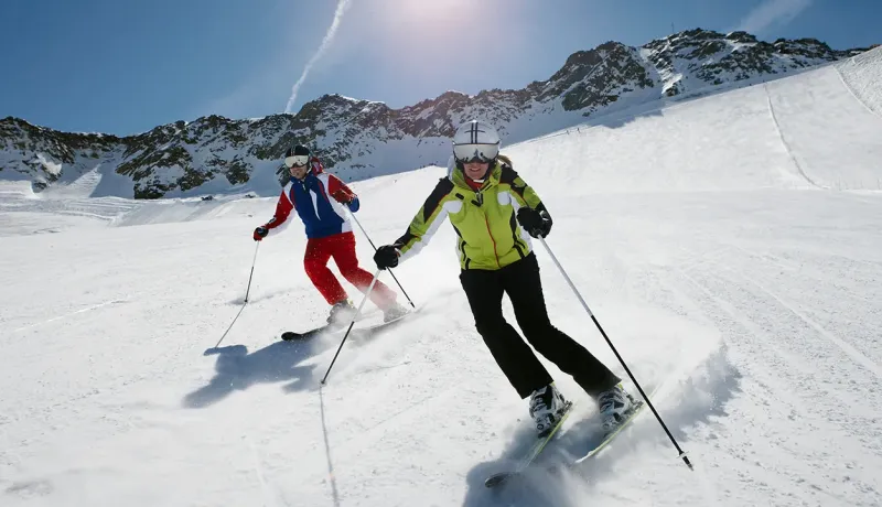 Un homme et une femme skient sur une piste enneigée sous un grand soleil.