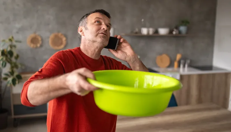 Homme inquiet au téléphone tenant une bassine pour recueillir une fuite d'eau du plafond.