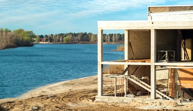 Chantier d'une maison moderne en béton sur un terrain sablonneux en bord de rive, zone potentiellement inondable.