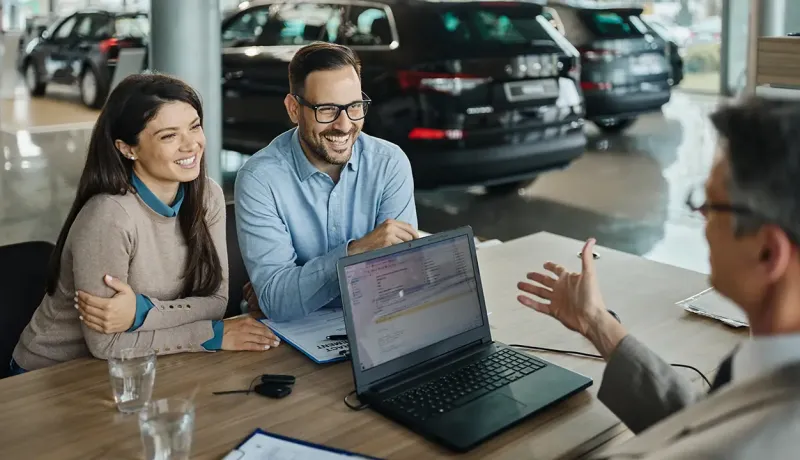 Dans une concession automobile, un couple consulte un vendeur à une table avec un ordinateur portable pour acheter un véhicule.