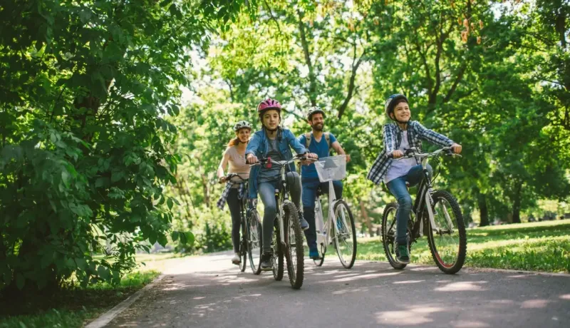Une famille de quatre personnes portant des casques circule à vélo sur un chemin ombragé au milieu d'un parc verdoyant.