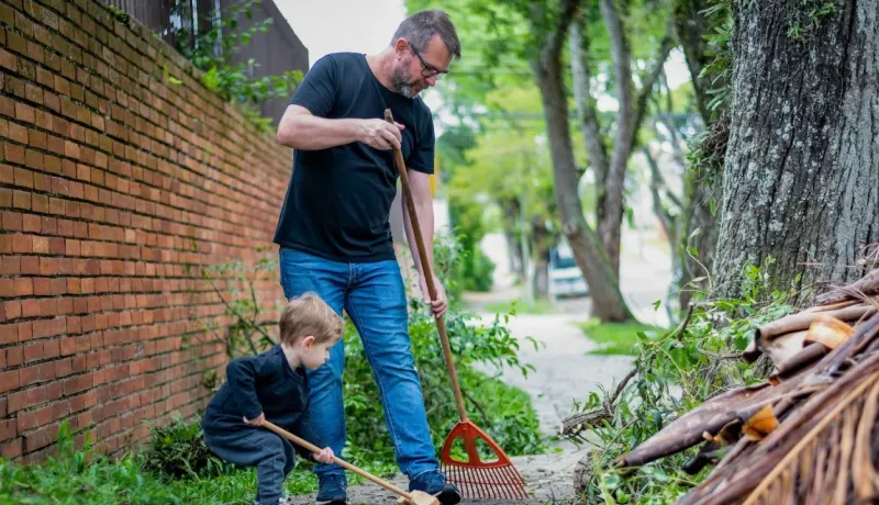 Un homme adulte et un jeune enfant ratissent ensemble les feuilles et les débris au bord d'une allée dans un environnement verdoyant.