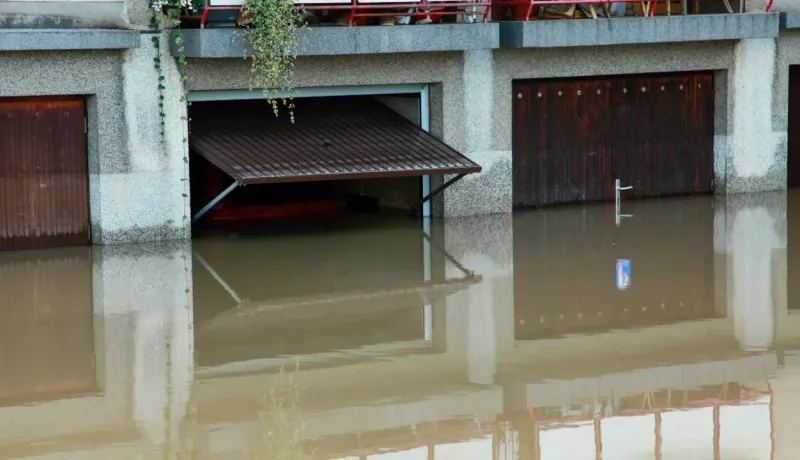 Des garages en pierre sont partiellement inondés par une eau boueuse, avec une porte entrouverte révélant une voiture submergée.