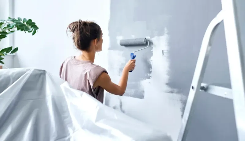 Femme de dos peignant un mur en gris avec un rouleau, protection en plastique au premier plan.