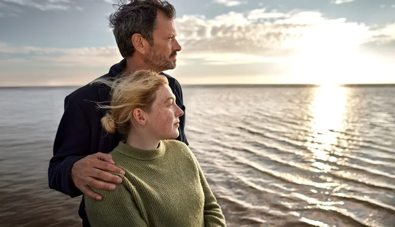 Un homme pose sa main sur l’épaule d’une jeune femme tandis qu’ils regardent l’horizon devant la mer.