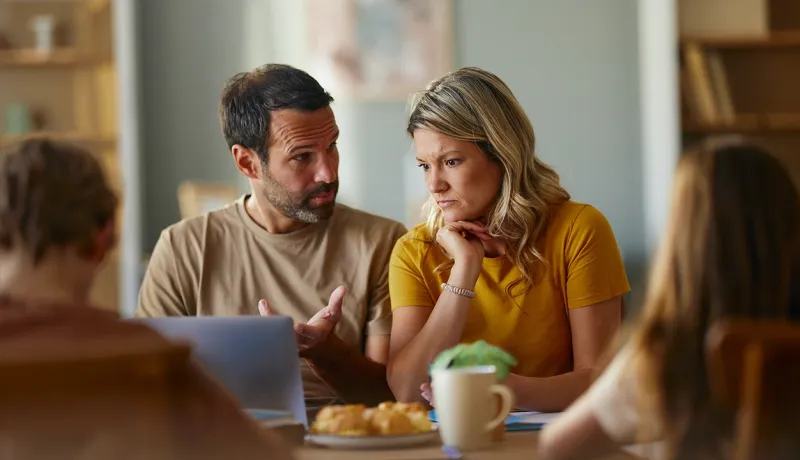 Un couple semblant préoccupé discute de démarches administratives devant un ordinateur portable, entouré de deux enfants dont on aperçoit le dos.