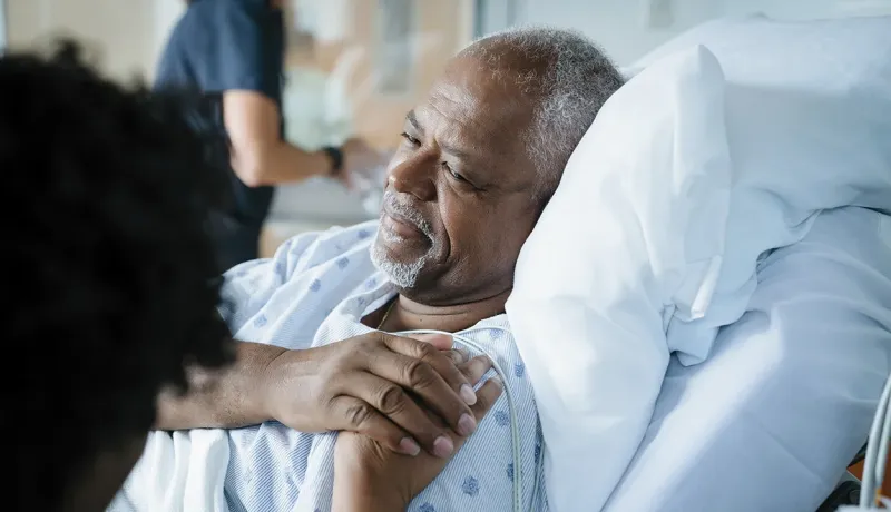 Un homme sur un lit d'hôpital, regardant sa femme avec un sourire complice et lui tenant la main.