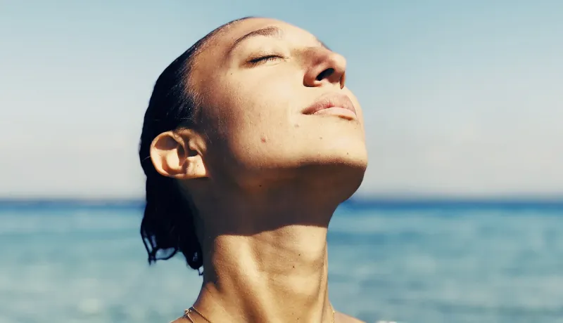 Femme au bord de la mer, visage tourné vers le soleil, yeux fermés, profitant d’un moment de bien-être en extérieur.