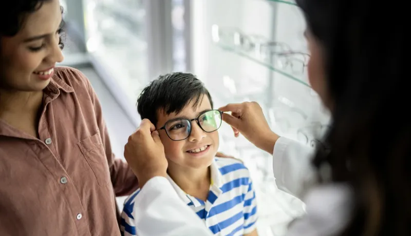 Une femme et son fils dans un magasin d'optique. L'opticien ajuste les nouvelles lunettes du jeune garçon, qui regarde, un grand sourire sur son visage.