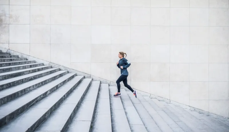 Une femme en tenue de sport court en montant de grands escaliers extérieurs en pierre devant un mur moderne quadrillé.