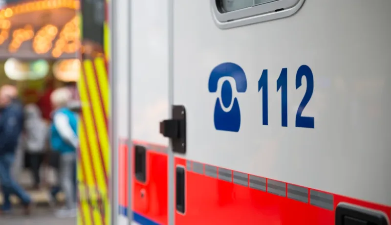 Gros plan sur le côté d'une ambulance avec le numéro d'urgence 112 écrit en bleu. À l'arrière-plan, la foule est floutée.