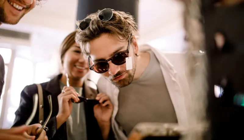 Un homme souriant essaye des lunettes de soleil. Ses amis sont derrière lui, l'air amusés. Ils se trouvent dans une boutique d'optique.