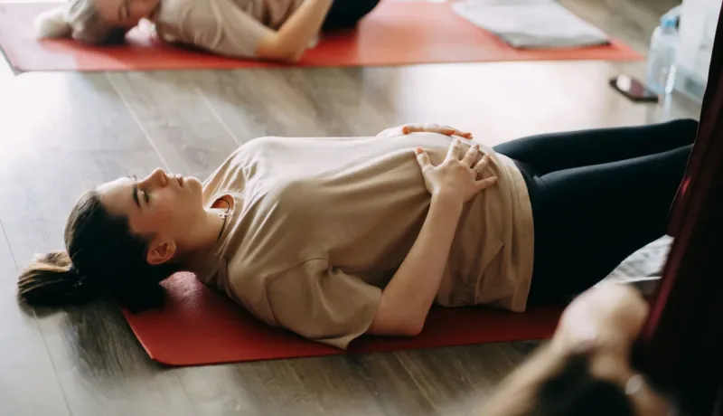 Une femme enceinte fait des exercices de respiration allongée sur un tapis de yoga rouge. Elle est dans un cours prénatal avec d'autres femmes.
