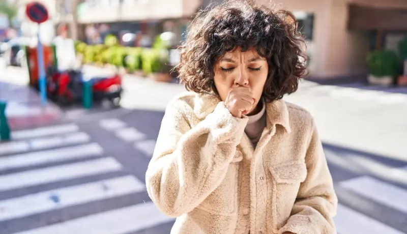 Une femme aux cheveux bouclés tousse ou éternue en pleine rue, les yeux fermés.