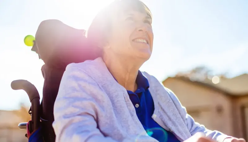Une personne souriante en fauteuil roulant, baignée de lumière solaire, portant un gilet gris et une chemise bleue, à l'extérieur.