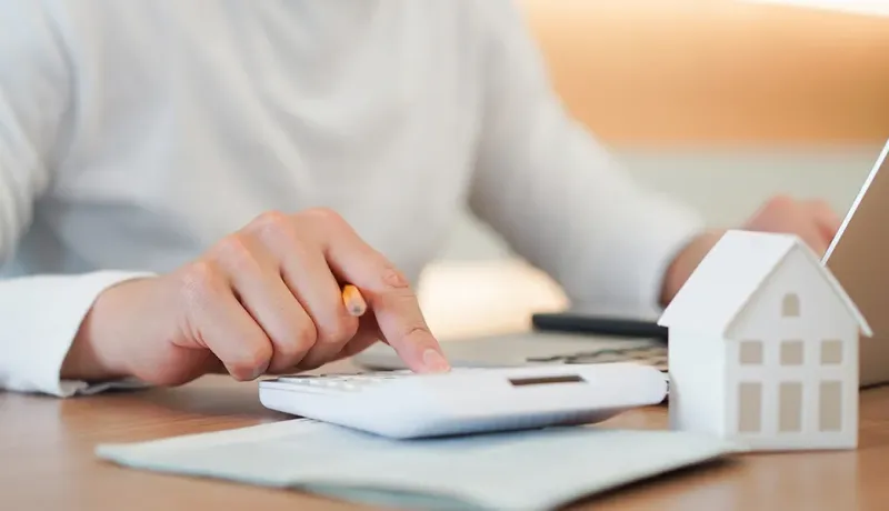 Un professionnel effectue des calculs sur une calculatrice et un ordinateur, avec une figurine de maison à côté.