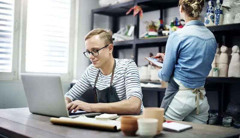 Un professionnel utilise un ordinateur portable pendant que sa collègue examine des produits artisanaux dans leur magasin.