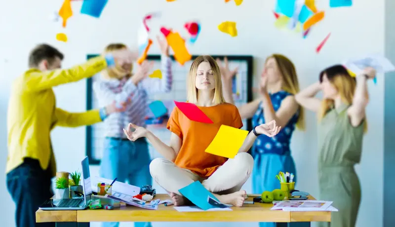 Une femme médite assise en tailleur sur un bureau au milieu du chaos et de collègues stressés en entreprise.