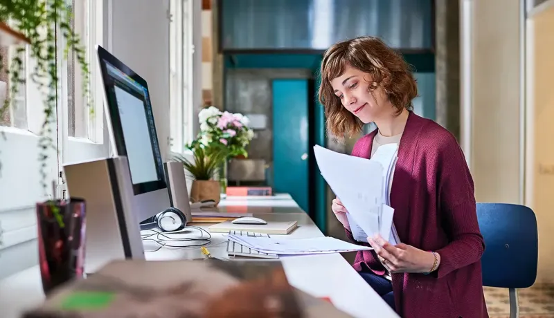 Une employée de bureau souriante assise à son poste de travail examine des documents administratifs papier.