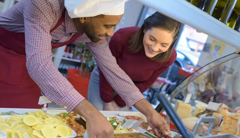 Un employé en toque et tablier sert une cliente souriante devant le comptoir d'une épicerie
