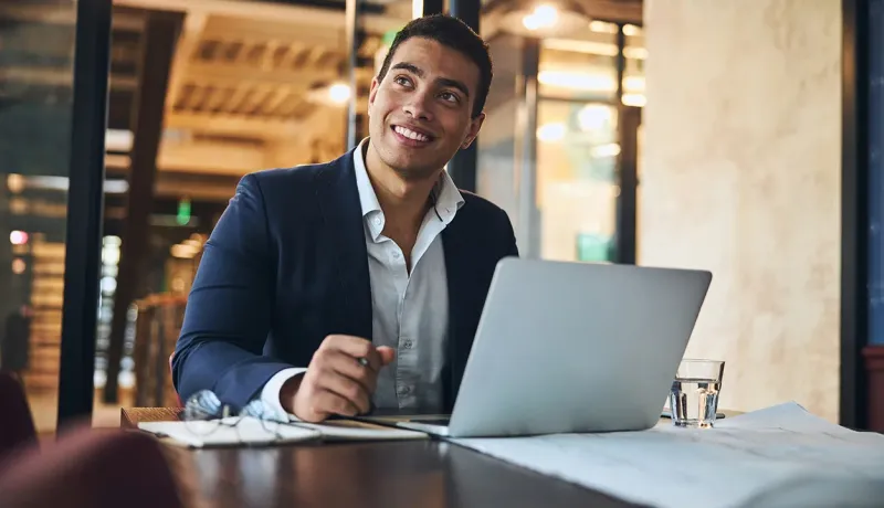Un homme en costume souriant et regardant au loin, assis à une table de bureau avec un ordinateur portable