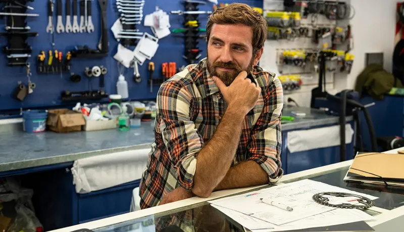 Un homme concentré, assis à une table dans un atelier, avec divers outils et équipements autour de lui.