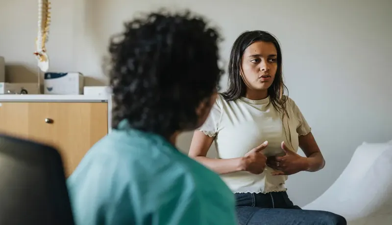Une patiente assise en consultation médicale montre et explique ses douleurs abdominales à un médecin.
