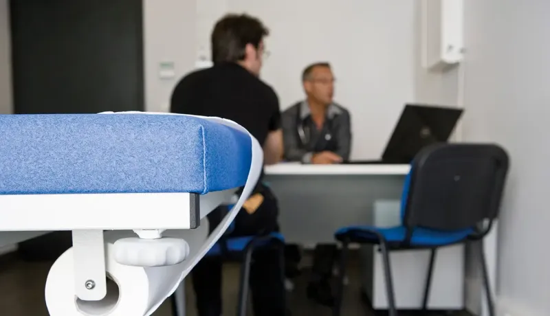Premier plan sur une table d’examen médicale bleue avec un médecin consultant un patient en arrière-plan.