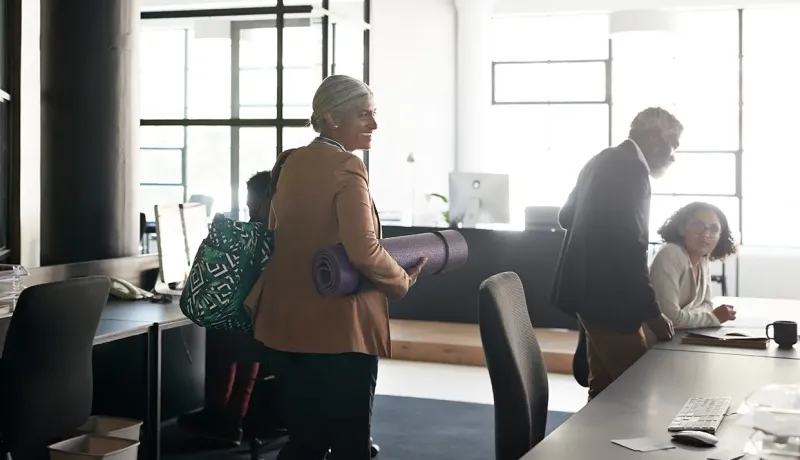 Une femme souriante traverse un bureau moderne en portant un tapis de yoga violet sous le bras.
