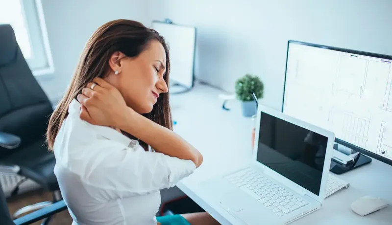 Une employée de bureau assise exprime une vive douleur au cou et aux cervicales devant son ordinateur portable.