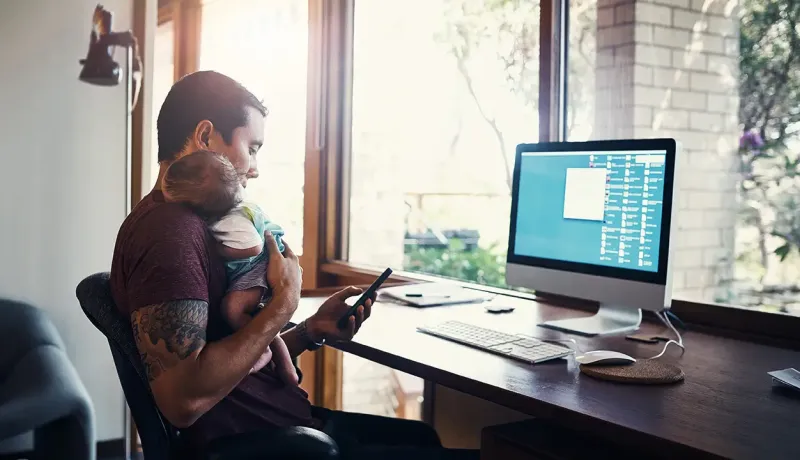 Homme en télétravail tenant son bébé dans les bras face à son ordinateur et son smartphone.