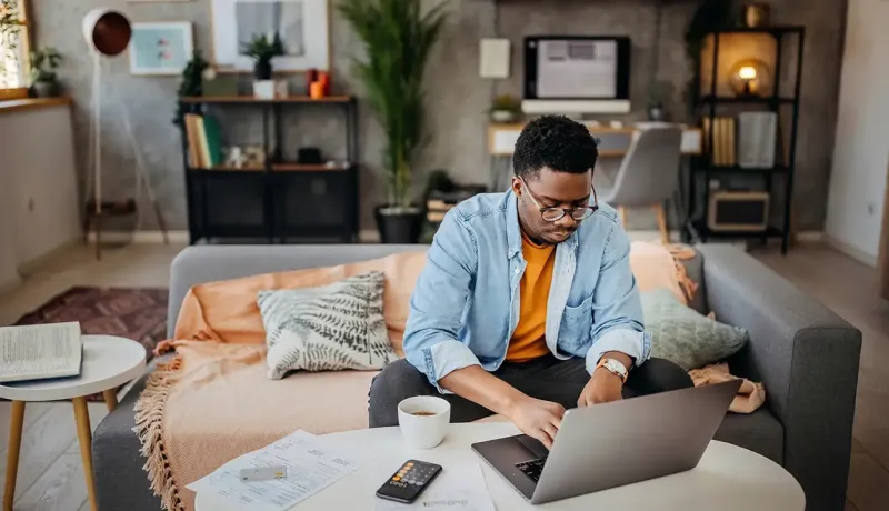 Un homme assis sur un canapé, travaille sur un ordinateur portable posé sur une table basse, dans un salon moderne.